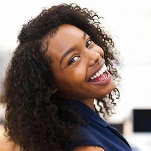 A close-up, cheerful portrait of a young Black woman with long, dark, curly hair and a nose piercing, smiling widely and looking over her shoulder.
