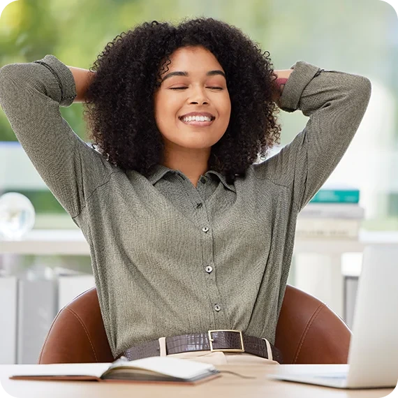 A young woman with curly brown hair sits at a desk with her hands behind her head, smiling and with her eyes closed in a moment of relaxation and stress relief in a modern office.