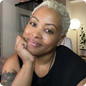 A close-up portrait of Kerrin St. Omer, a Black woman with short, platinum blonde hair, smiling softly with her chin resting on her hand, taken indoors.