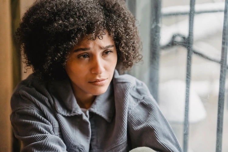A close-up portrait of a woman with curly brown hair looking down and away with a troubled expression while sitting near a window with a snowy view outside.